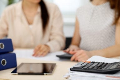 Two female accountants counting on calculator income for tax form completion hands closeup. Internal Revenue Service inspector checking financial document. Planning budget