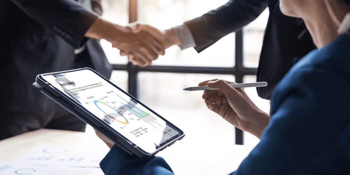 Going Through M&A? Make These Technology Decisions During Due Diligence - business people shake hands over a deal as a third person makes notes on a tablet